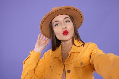 Beautiful young woman blowing kiss while taking selfie on purple background