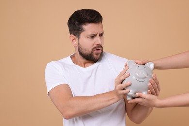Photo of Woman taking piggy bank from emotional man on beige background. Be careful - fraud