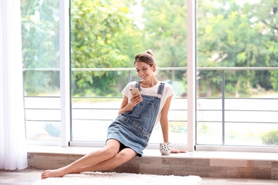 Photo of Young woman with mobile phone sitting near window at home
