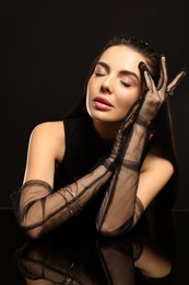Beautiful young woman in evening gloves at black glass table against dark background