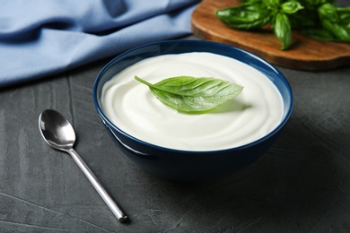 Photo of Bowl of fresh sour cream with basil and spoon on grey table