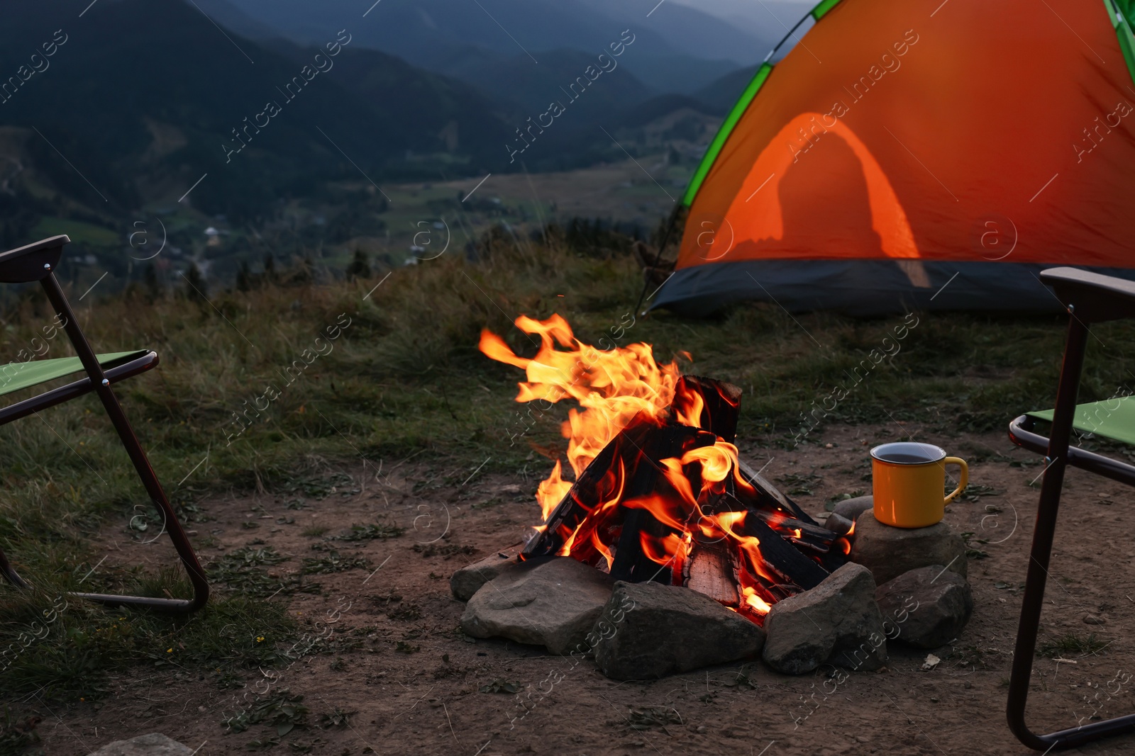 Photo of Metal mug with drink and beautiful bonfire near camping tent in mountains