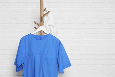 Photo of Blue medical uniform hanging on rack near white brick wall. Space for text