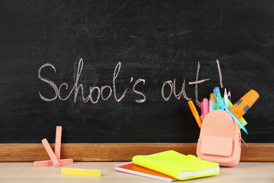 Photo of Pieces of color chalk and stationery on table near blackboard with text School's Out. Summer holidays