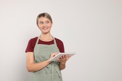 Photo of Beautiful young woman in clean apron with tablet on light grey background. Space for text