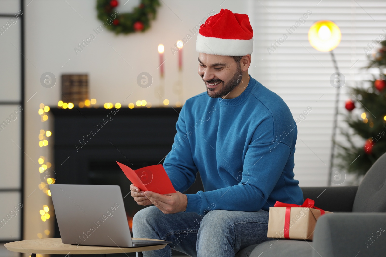 Photo of Celebrating Christmas online with exchanged by mail presents. Smiling man in Santa hat reading greeting card during video call on laptop at home