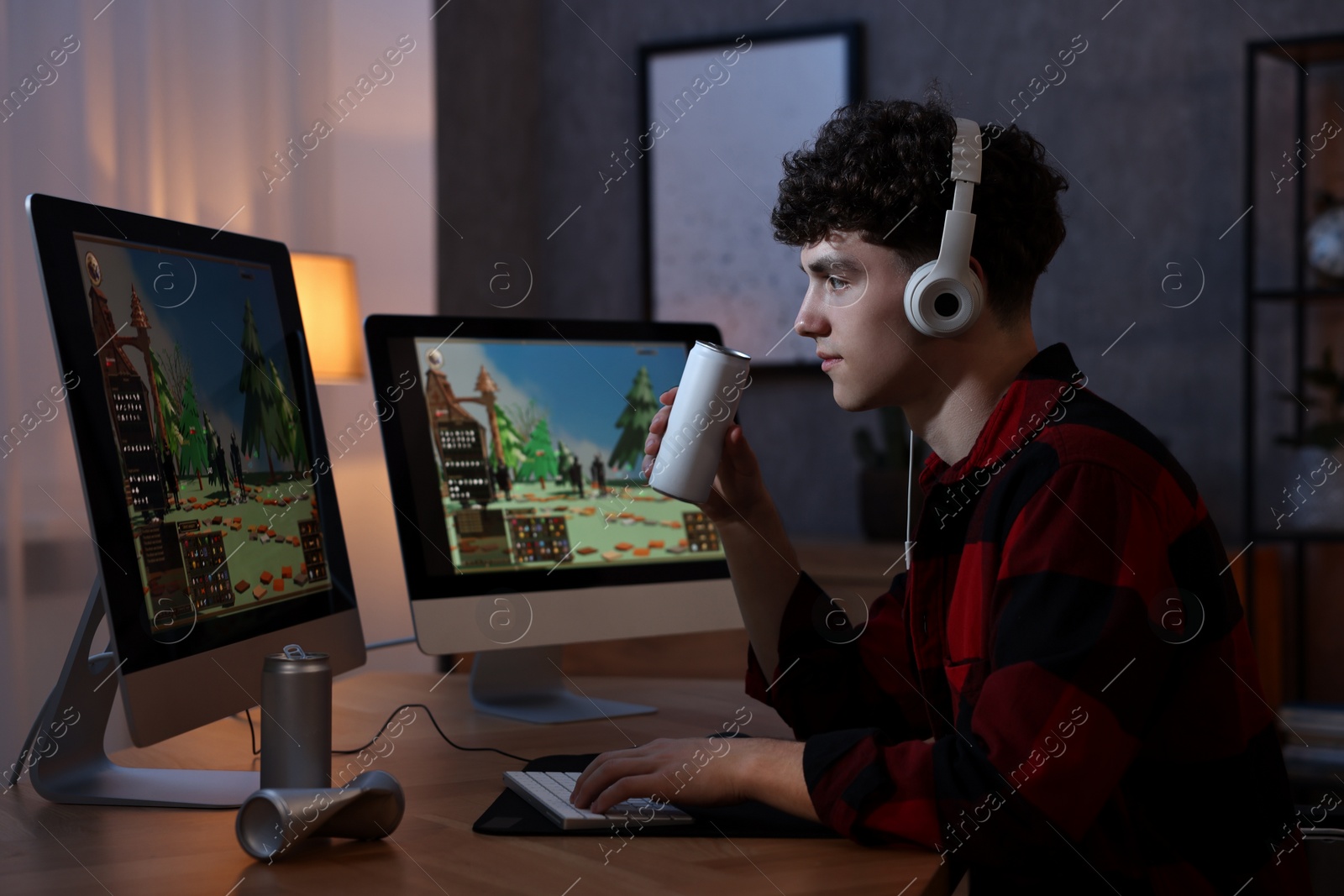 Photo of Young man with energy drink and headphones playing video game at wooden desk indoors