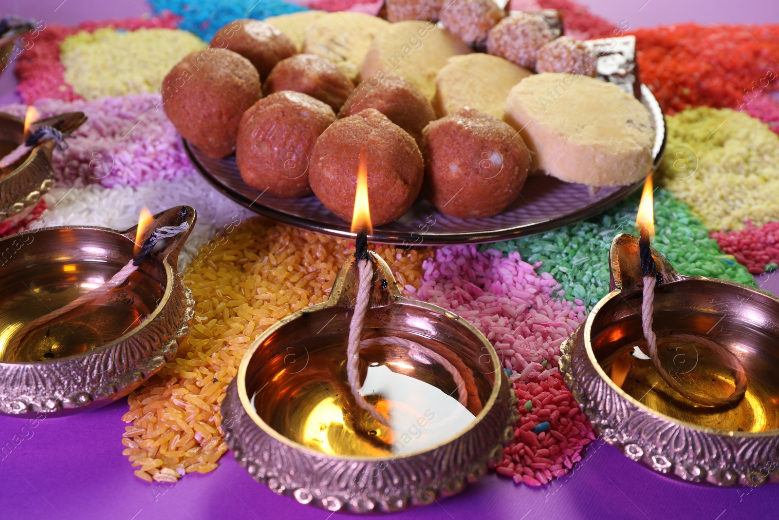 Photo of Diwali celebration. Tasty Indian sweets, diya lamps and colorful rangoli on violet table, closeup