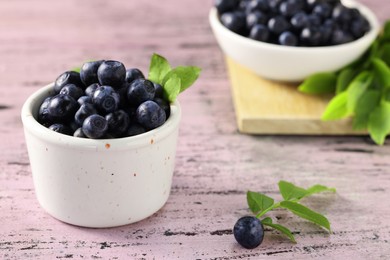 Tasty fresh bilberries with green leaves in bowls on old pink wooden table, closeup. Space for text