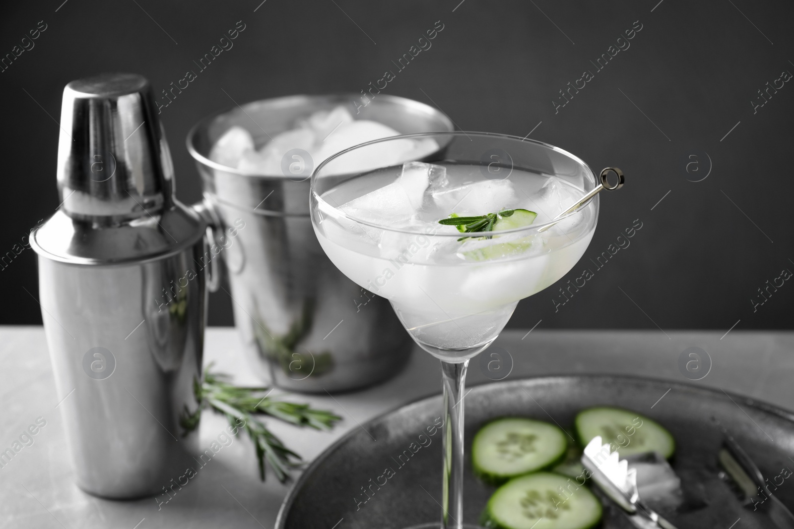 Photo of Glass of martini with cucumber, shaker and ice bucket on table against dark background. Space for text
