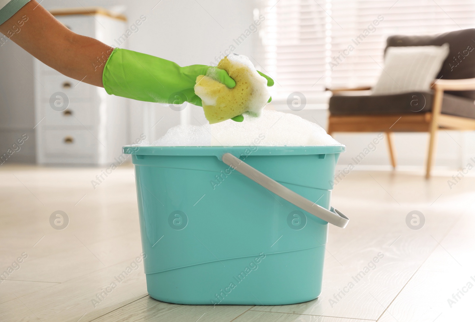 Photo of Woman holding sponge with foam over bucket indoors, closeup. Cleaning supplies