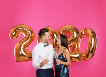 Photo of Happy young couple with glasses of champagne near golden 2020 balloons on pink background. New Year celebration