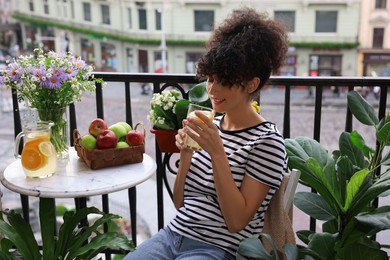 Young woman with glass of refreshing drink near beautiful houseplants on balcony