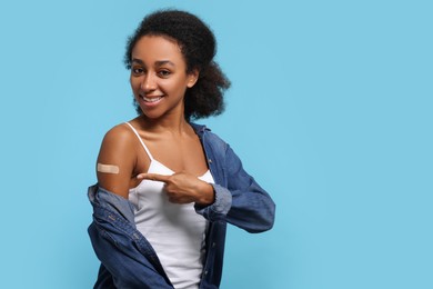 Photo of Happy young woman pointing at adhesive bandage on her arm after vaccination against light blue background. Space for text