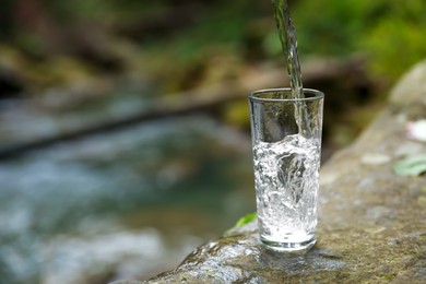 Photo of Fresh water pouring into glass on stone near stream. Space for text