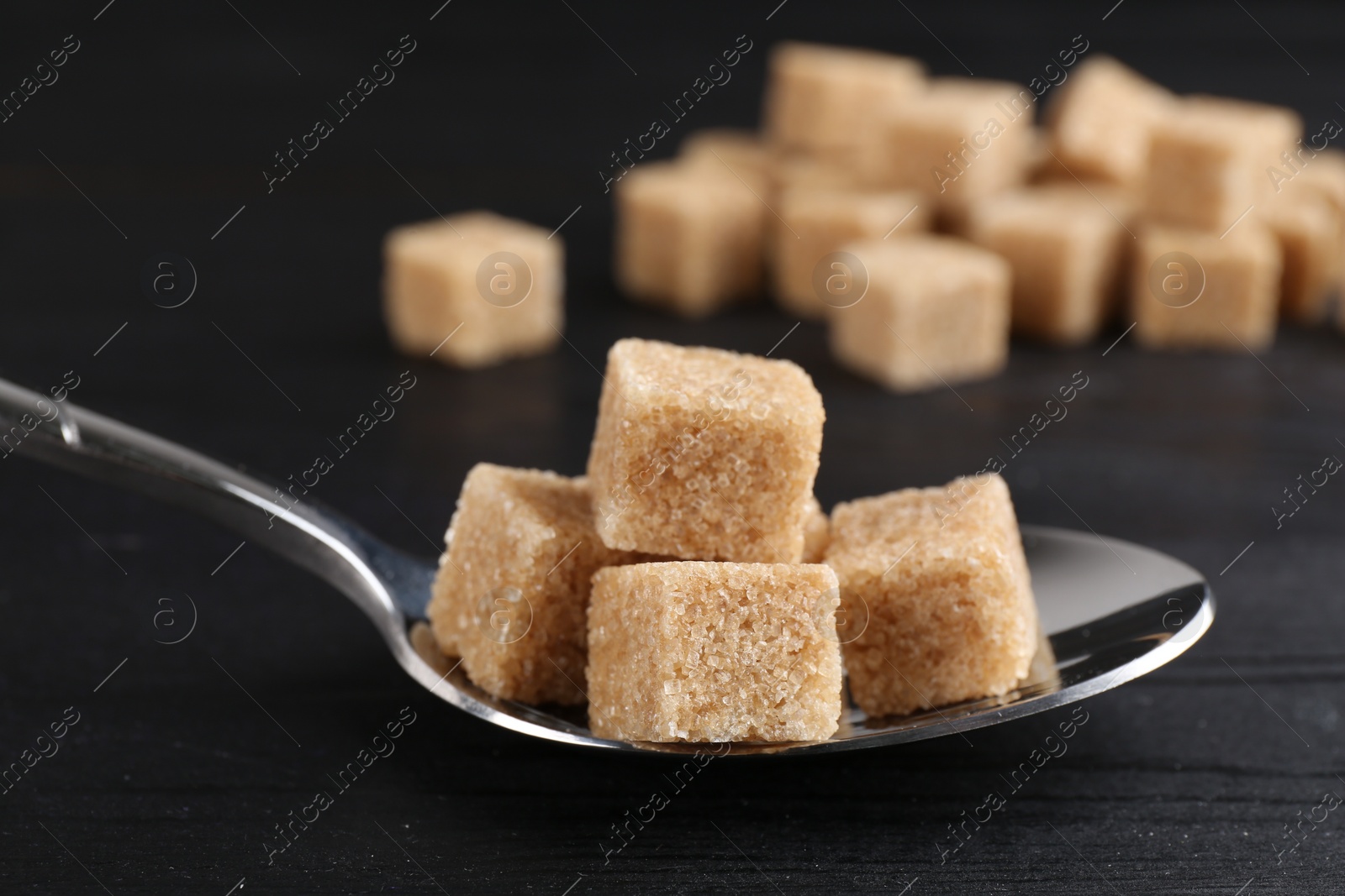 Photo of Brown sugar cubes in spoon on black wooden table, closeup