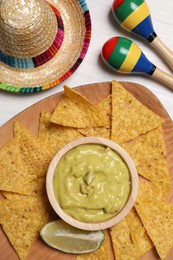 Delicious guacamole with nachos chips, Mexican sombrero hat and maracas on white wooden table, flat lay