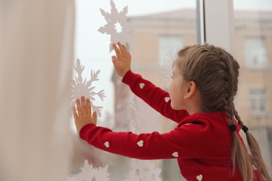 Cute little girl decorating window with paper snowflake indoors