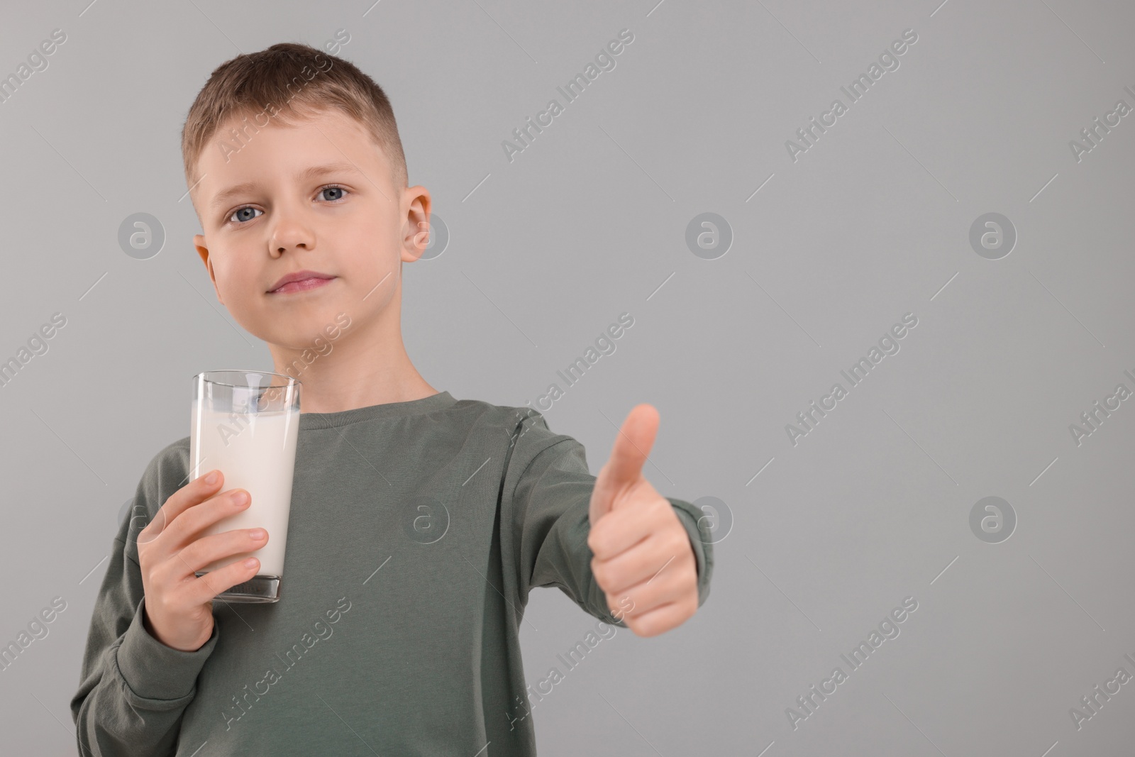 Photo of Cute boy with glass of fresh milk showing thumb up on light grey background, space for text