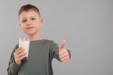 Cute boy with glass of fresh milk showing thumb up on light grey background, space for text
