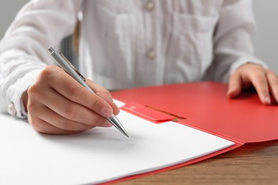 Photo of Woman writing on sheet of paper in red folder at wooden table in office, closeup