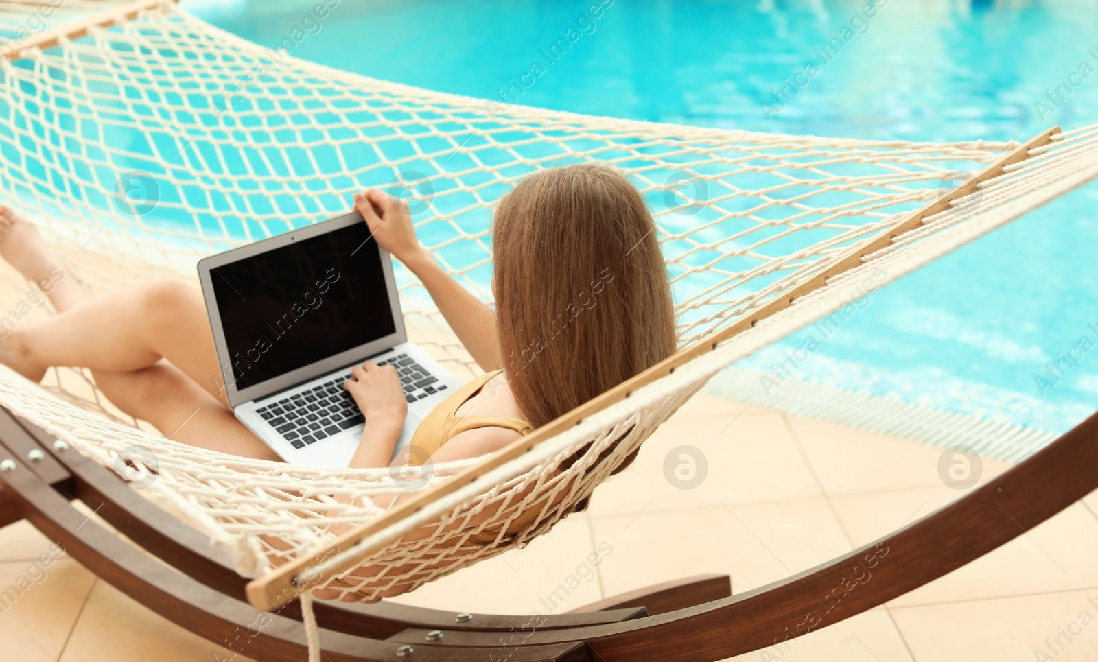 Photo of Young woman with laptop resting in hammock near pool