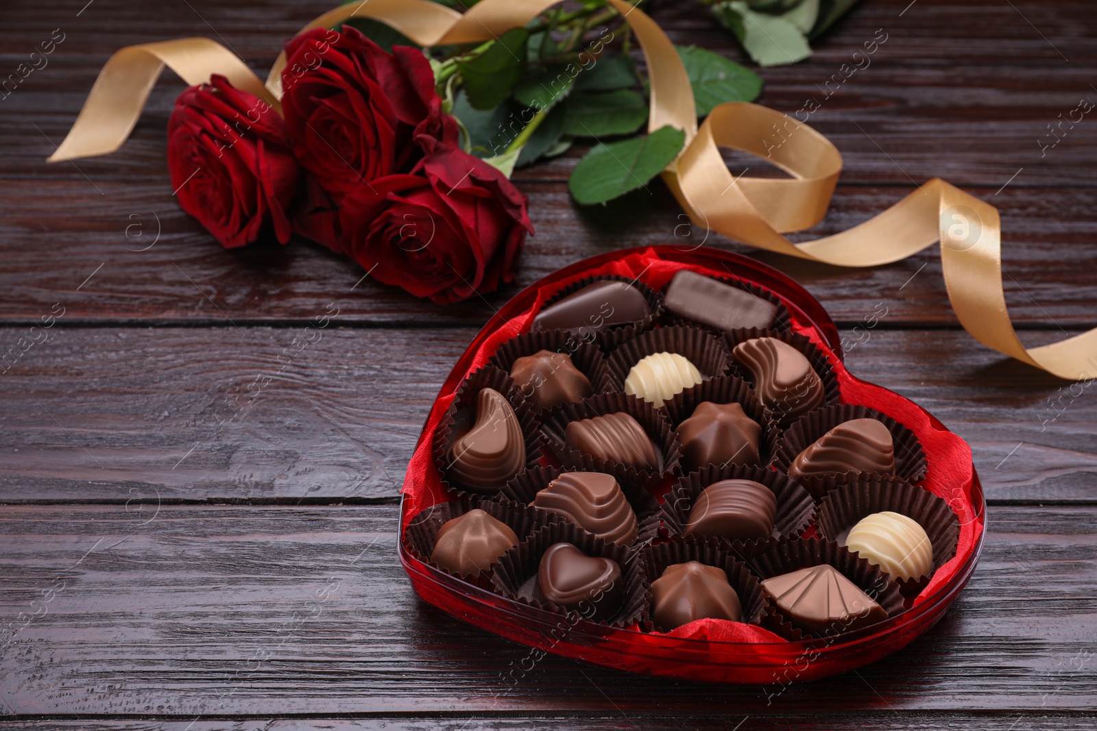 Photo of Heart shaped box with delicious chocolate candies, roses and ribbon on wooden table. Space for text