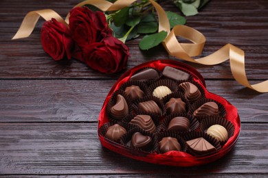 Photo of Heart shaped box with delicious chocolate candies, roses and ribbon on wooden table. Space for text