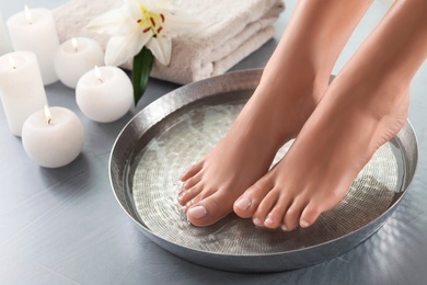 Closeup view of woman soaking her feet in dish with water on grey floor. Spa treatment