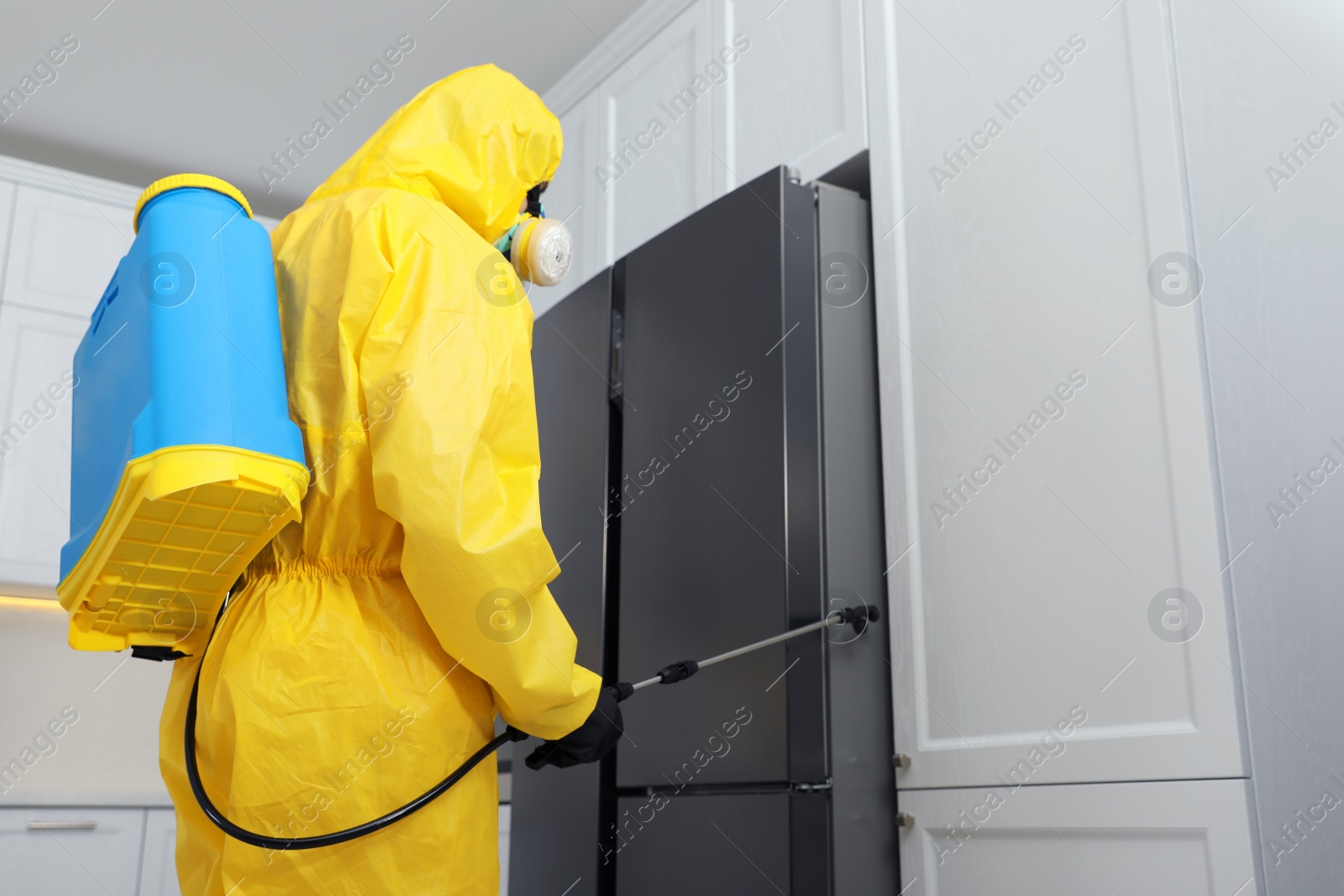 Photo of Pest control worker in protective suit spraying insecticide near refrigerator indoors