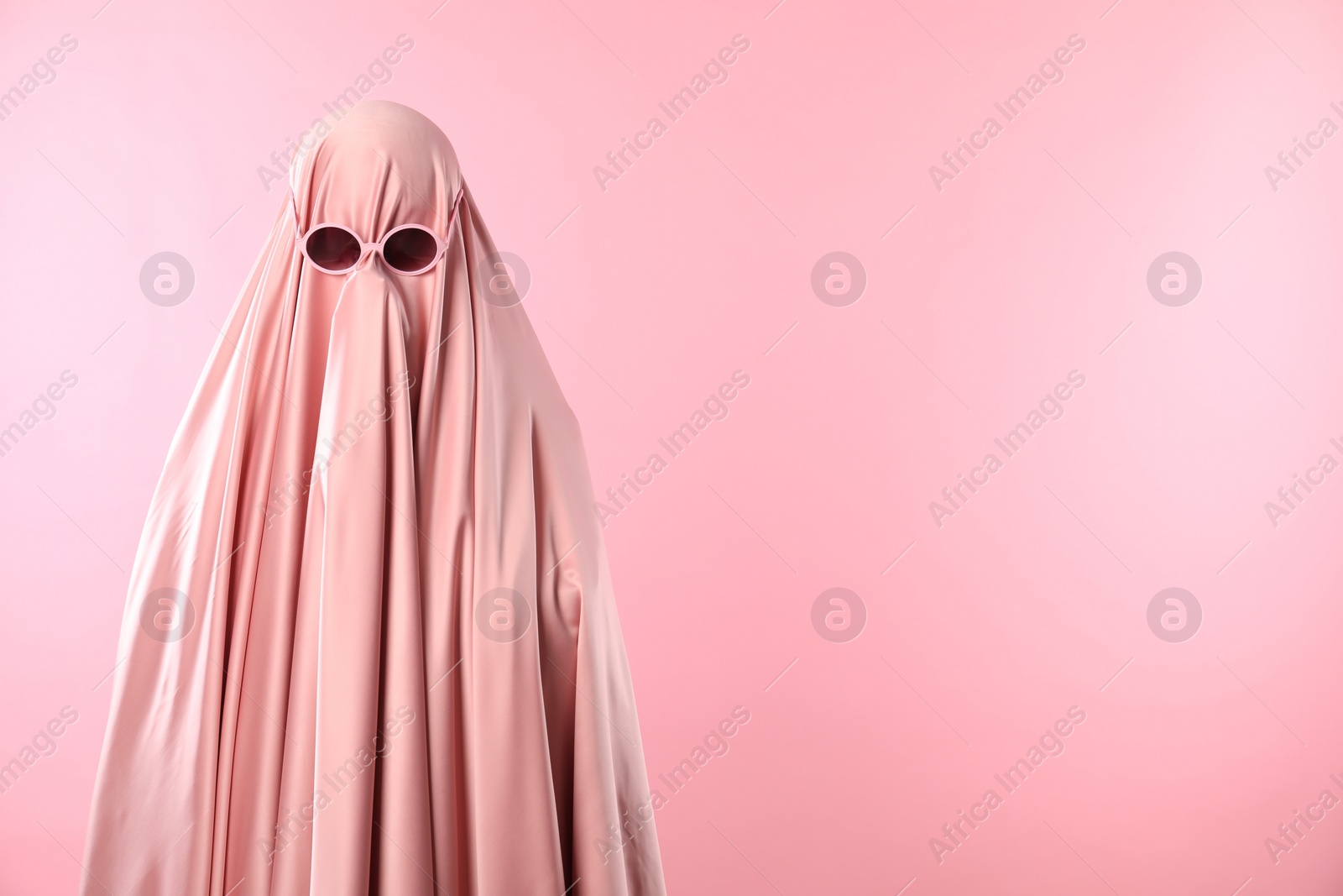 Photo of Glamorous ghost. Woman in sheet with sunglasses on pink background, space for text