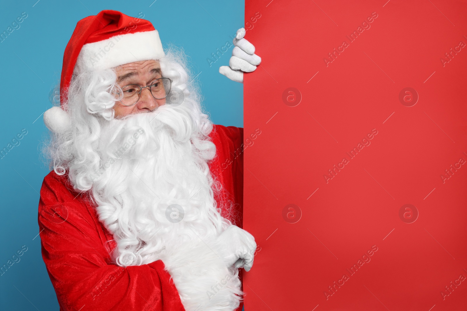 Photo of Santa Claus holding blank red poster on light blue background, space for text