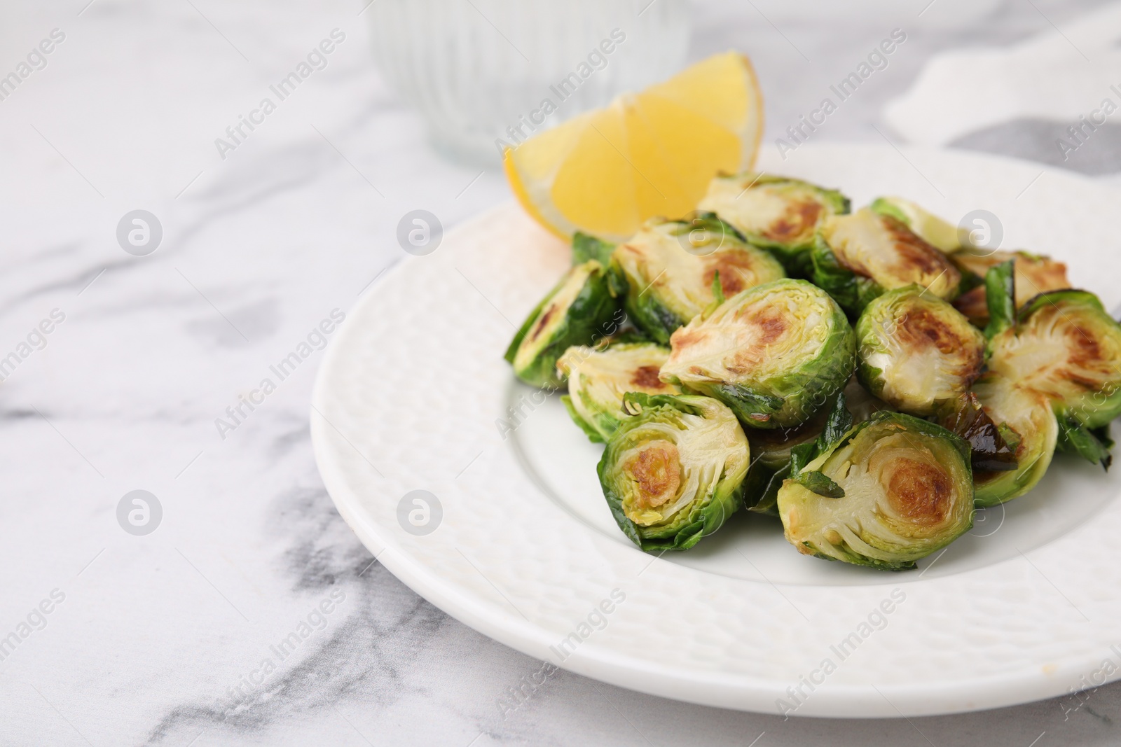 Photo of Delicious roasted Brussels sprouts and slice of lemon on white marble table, closeup. Space for text