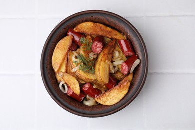 Photo of Delicious baked potato with thin dry smoked sausages, onion and dill in bowl on white tiled table, top view