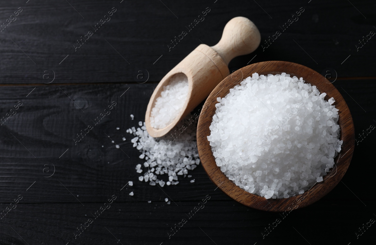 Photo of Organic salt in bowl and scoop on black wooden table, top view. Space for text