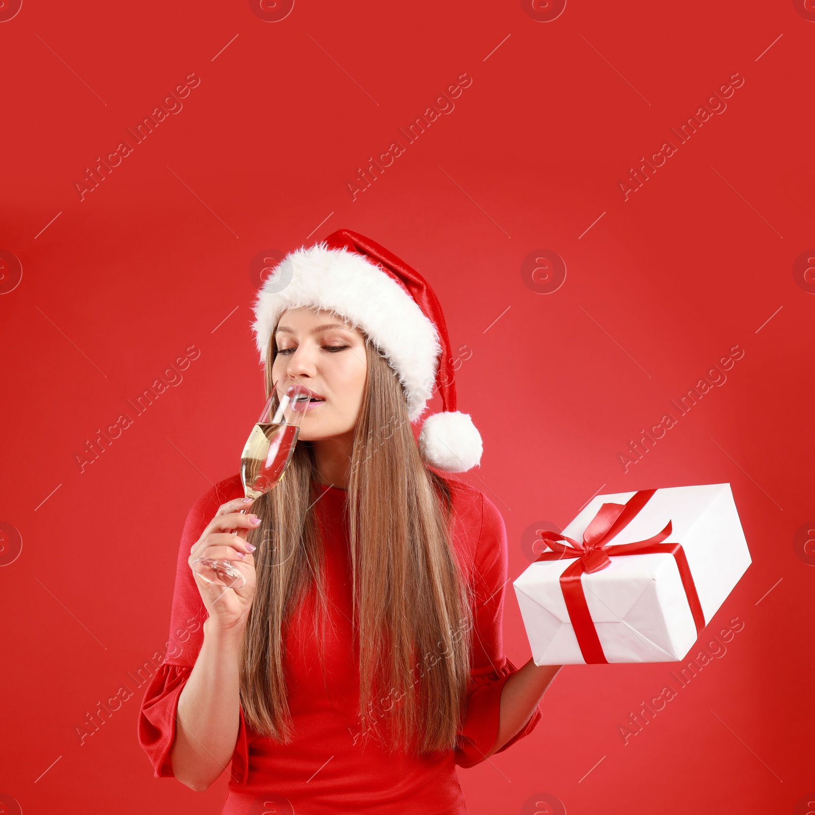 Photo of Young woman in Santa hat with Christmas gift and glass of champagne on red background