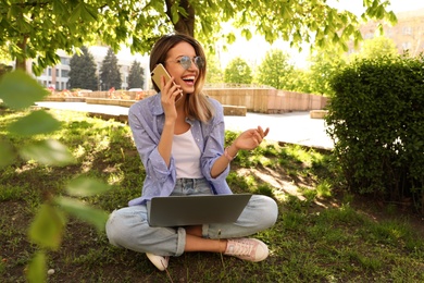 Image of Happy young woman with laptop talking on phone in park 