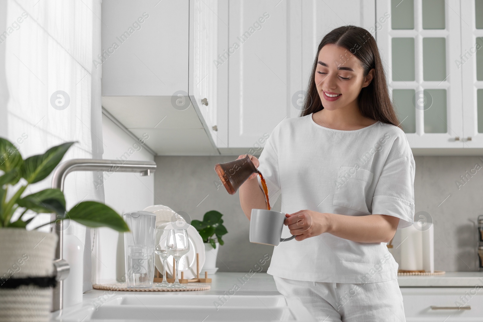 Photo of Beautiful woman pouring coffee from jezve into cup in kitchen. Lazy morning