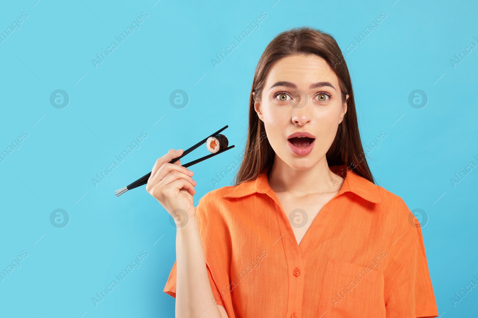 Photo of Emotional young woman holding sushi roll with chopsticks on light blue background