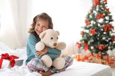 Photo of Cute little child with toy bear and Christmas gift box sitting on bed at home