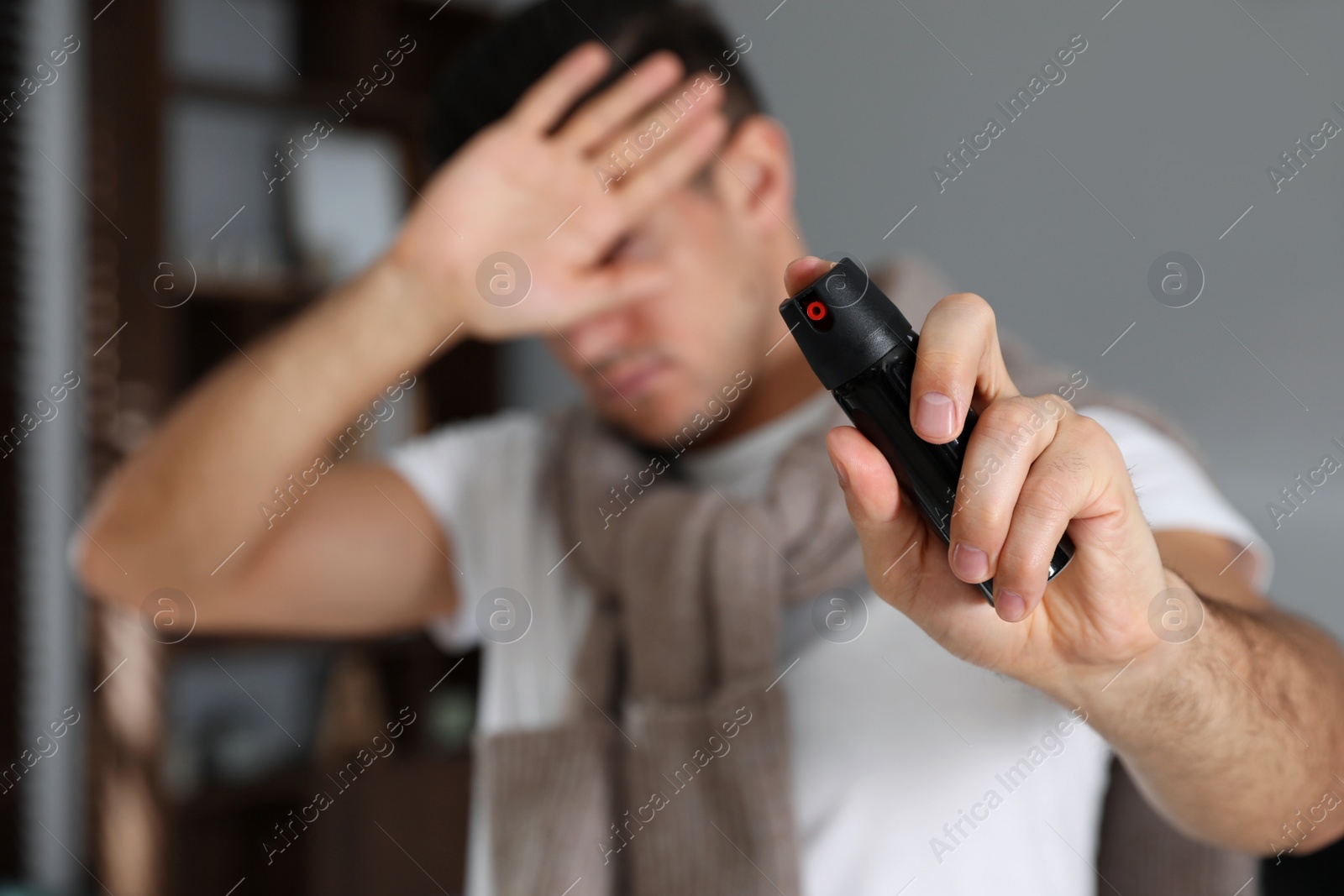 Photo of Man covering eyes with hand and using pepper spray indoors, focus on canister