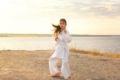 Photo of Cute little girl in kimono practicing karate near river on sunny day