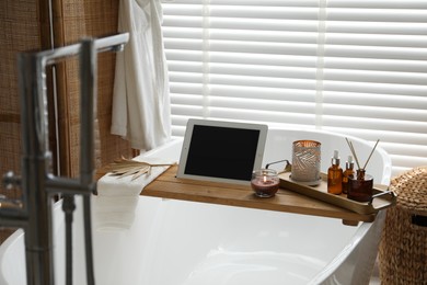 Photo of Wooden tray with tablet, cosmetic products and burning candles on bath tub in bathroom