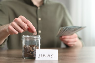 Financial savings. Man putting coin into glass jar at wooden table, closeup