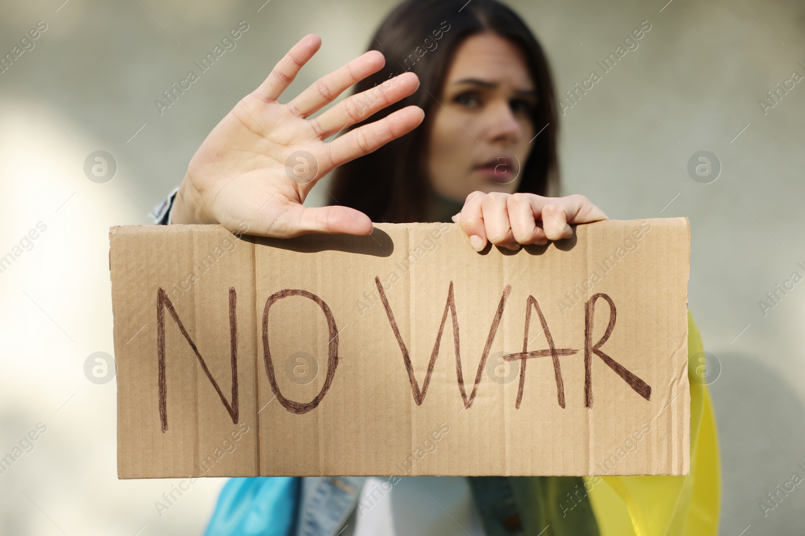 Photo of Sad woman holding poster with words No War and showing stop gesture near light wall, focus on hands