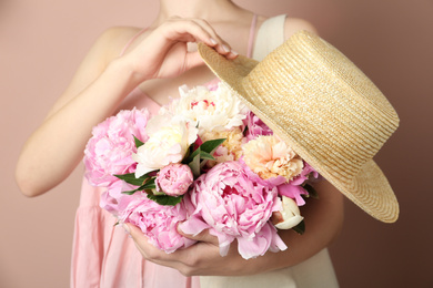Photo of Woman with bouquet of beautiful peonies in bag and hat on beige background, closeup