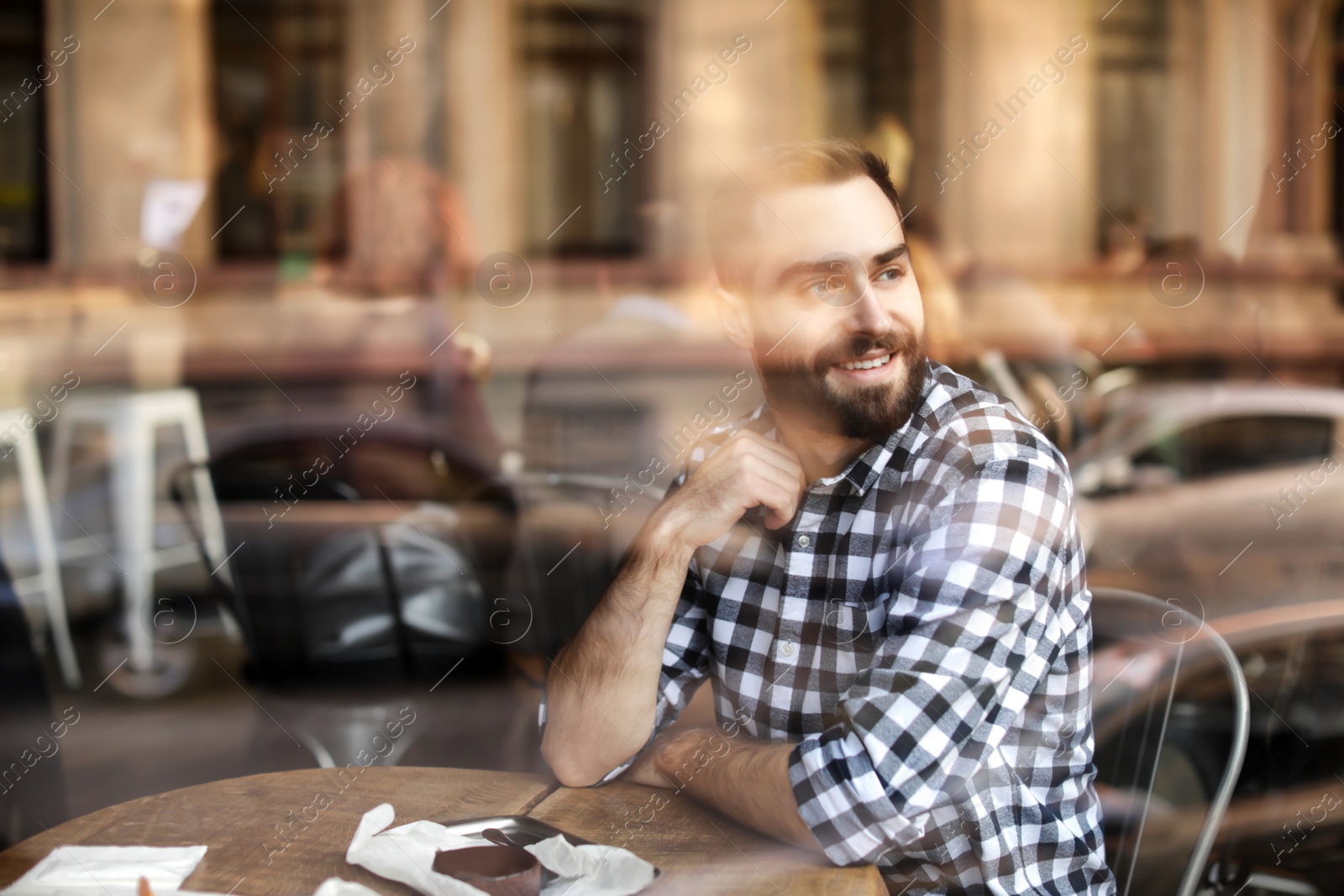 Photo of Handsome young man sitting at table in cafe, view from outdoors through window