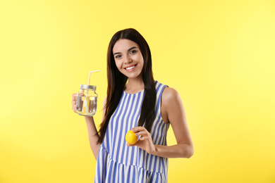 Beautiful young woman with tasty lemon water and fresh fruit on yellow background