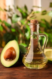 Photo of Glass jugcooking oil and fresh avocados on wooden table against blurred green background