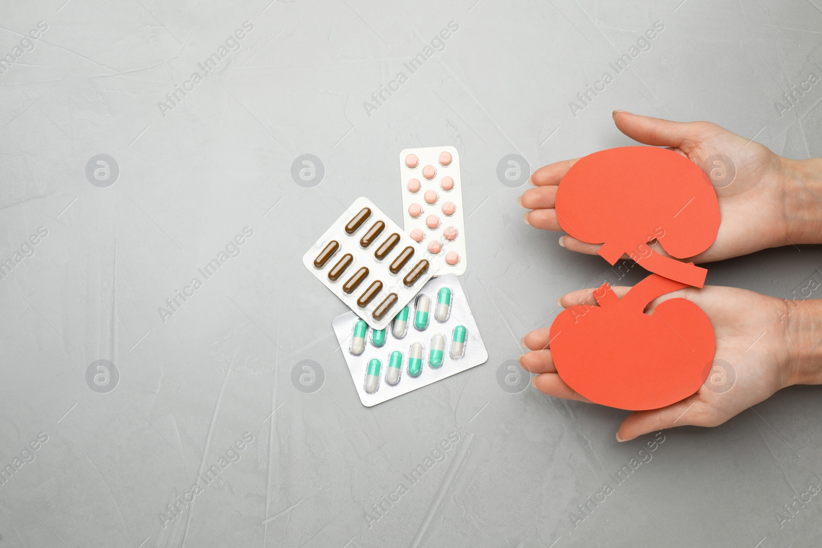 Photo of Woman holding paper cutout of kidneys near pills at grey table, top view. Space for text